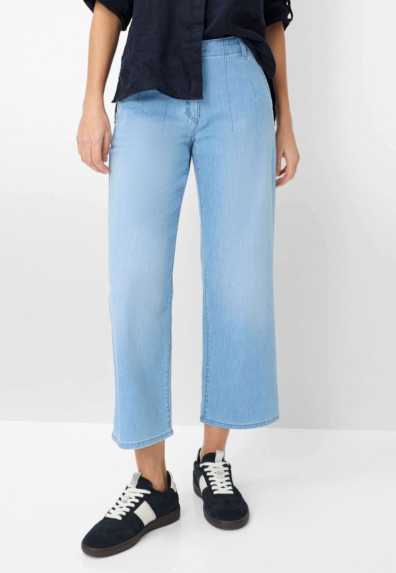 Person iført lyseblå cropped vidde bukser, sorte og hvide sneakers samt en sort langærmet skjorte med ærmerne rullet op.