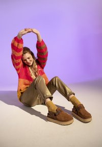 Zoccoli in suede marrone chunky con suola beige, indossati con pantaloni metallici color oliva e calzini a coste color senape, su uno sfondo viola.
