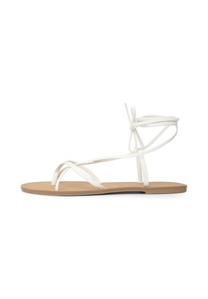 Sandale à semelle plate couleur beige clair avec fines lanières blanches qui croisent les orteils et entourent la cheville, nouées en un nœud à l'arrière.