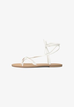 Sandale à semelle plate couleur beige clair avec fines lanières blanches qui croisent les orteils et entourent la cheville, nouées en un nœud à l'arrière.