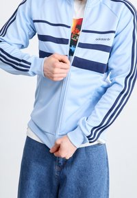 Ljusblå zip-jacka med marinblå ränder över bröstet och ärmarna; mjukt tyg och ribbad mudd, kombinerad med denimbyxor.