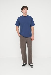 Μπλε ναυτικό κοντομάνικο t-shirt με στρογγυλή λαιμόκοψη, συνδυασμένο με καφέ παντελόνι ίσιας γραμμής με εμφανείς ραφές και μαύρα αθλητικά παπούτσια.