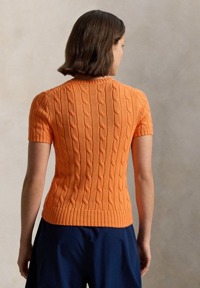 Γυναίκα με κοντά καστανά μαλλιά, φορώντας εφαρμοστή πορτοκαλί κοντομάνικη μπλούζα πλεκτή με σχέδιο κοτσίδα και μπλε σκούρο παντελόνι, γυρισμένη πλάτη σε ουδέτερο φόντο.