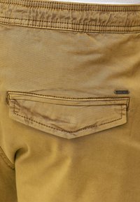 Pantalon kaki avec une poche arrière dotée d'un rabat pointu. Texture lisse avec des coutures visibles et une étiquette de marque sur la poche.