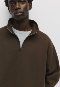 Hnedá mikina na zips s vysokým golierom, vyrobená z jemného materiálu. Má strieborný zips a je oblečená cez svetlo šedú košeľu.