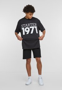 Crna oversized majica s bijelim printom "STARTER 1971", uparena s crnim kratkim hlačama i bijelim tenisicama na neutralnoj pozadini.
