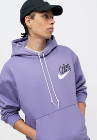 Fialová mikina vyrobená z jemného materiálu, so bielym logom Nike a šnúrkou s kovovými koncami. Obsahuje predné vrecko a rebrované manžety.