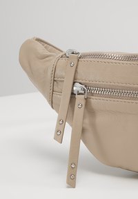 Sac en cuir beige avec deux fermetures éclair métalliques argentées et longues languettes en cuir avec des clous métalliques sur une surface blanche.