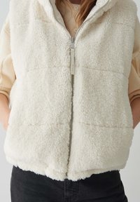 Vrouw in een crèmekleurige, donzige zip-up vest over een lichtbeige trui, met haar handen in de vestzakken en zwarte broek.