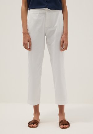 Personne portant un pantalon court blanc taille haute, un haut bleu marine boutonné et des sandales plates tressées marron, debout sur un fond clair uni.