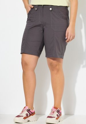 Person, die dunkelgraue knielange Shorts und pink-weiße-braune Sneakers trägt, steht vor einer hellen Wand.