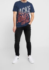 Muž oblečený v námořnickém modrém tričku s grafikou "Jack & Jones 1990", černých úzkých džínách a bílých teniskách Nike, stojí s jednou rukou v kapse.