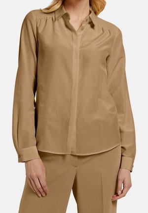 Blusa a maniche lunghe di colore beige, realizzata in tessuto morbido, con chiusura a bottoni davanti, colletto morbido e dettagli di arricciatura discreti sulle spalle.