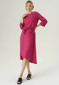 Abito fucsia con maniche a tre quarti, vita cintrata e orlo asimmetrico. Tessuto liscio, silhouette semplice, indossato con tacchi open-toe neri.