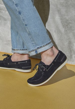 Granatowe zamszowe mokasyny żeglarskie z białymi sznurówkami i detalami przeszyć; lekkie podeszwy; noszone z podwiniętymi jasnoniebieskimi dżinsami na żółtej podłodze.