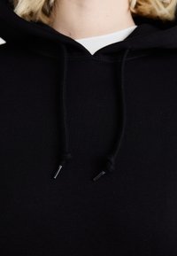 Felpa nera con cappuccio realizzata in tessuto morbido, dotata di un cordino con punte metalliche e una vestibilità comoda. I dettagli includono un colletto interno bianco.