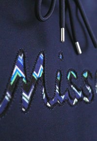 Tecido azul marinho com superfície texturizada, apresentando "Miss" bordado num padrão geométrico multicolorido. Cordão com pontas metálicas presente.