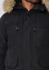 Parka noire avec capuche en fourrure, fermeture éclair et cordons de serrage. Comprend deux grandes poches avant et une texture lisse.