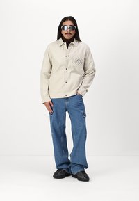 Beige button-up jacket with a logo on the chest pocket, paired with loose blue denim pants and black textured sneakers.