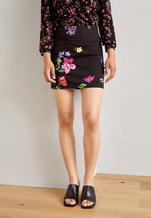 Mujer vistiendo una mini falda negra con estampado floral, combinada con una blusa negra de manga larga con pequeñas flores de colores y mules negros con tacón y punta abierta.