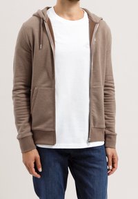 Man med en taupe zip-up huvtröja över en vit t-shirt och blå jeans, som står mot en ljus, enkel bakgrund.