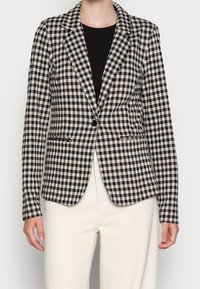 Blazer à carreaux noir et blanc avec un col cranté, une fermeture à un bouton et deux poches avant, associé à un pantalon de couleur crème.