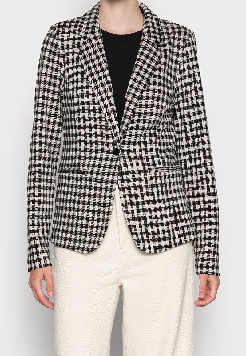 Blazer à carreaux noir et blanc avec un col cranté, une fermeture à un bouton et deux poches avant, associé à un pantalon de couleur crème.