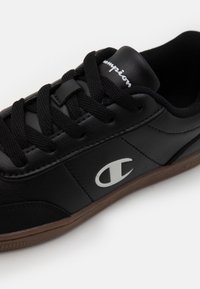 Baskets basses noires avec lacets noirs, logo Champion blanc sur le côté et semelle en caoutchouc marron, présentées sur un fond blanc.