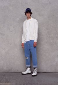 Giacca bomber bianca su una maglietta bianca, abbinata a jeans strappati azzurri chiari e stivali da combattimento bianchi. Un cappellino blu completa il look.