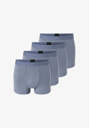 Hemdloser Mann in grauen Boxershorts steht mit erhobenen Fäusten, schaut aufmerksam zur Seite vor einem schlichten Hintergrund.