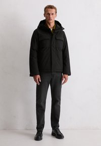 Zwarte gevoerde jas met capuchon, voorzien van twee borstzakken en een ritssluiting, gecombineerd met donkere jeans en zwarte schoenen.