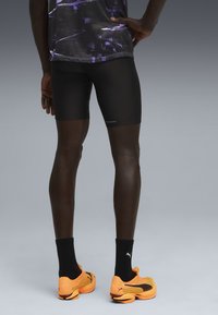 Sorte atletiske shorts, orange Puma løbesko med sorte accenter og sorte sokker. Glat tekstur, tætsiddende design, velegnet til sport.