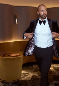 Giacca da smoking nera in velluto con rivestimento a motivo floreale, camicia bianca con pence, papillon nero e pantaloni neri in un ambiente moderno.