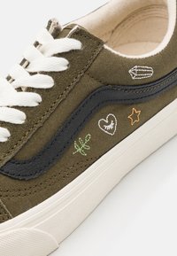Zapatillas de lona verde oliva con cordones blancos, una franja de cuero negro y detalles bordados que incluyen un corazón, una estrella y un diseño de planta.