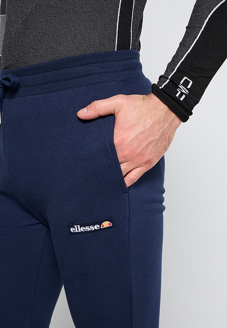 Dunkelblaue Jogginghose mit einem Kordelzugbund, ausgestattet mit Seitentaschen und einem gestickten ellesse-Logo-Patch am linken Oberschenkel. Weicher Stoff.