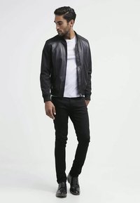 Uomo con giacca di pelle nera, t-shirt bianca, pantaloni neri e scarpe eleganti nere, in piedi con le mani in tasca mentre guarda a sinistra.