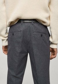 Pantaloni gri cărbune cu o textură fină, având buzunare posteriori cu nasturi, purtați cu un pulover cremos.