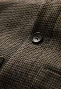 Brunt houndstooth-stof med en fin tekstur, med en sort knap med fire huller syet på beklædningen. Nærbillede af materialet.