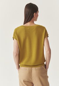 Femme aux longs cheveux noirs portant un haut en tricot à manches courtes jaune moutarde et un pantalon beige, se tenant avec les mains dans les poches, de dos.