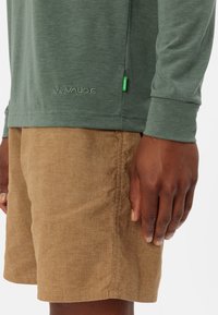 Camicia a maniche lunghe verde bosco con un logo sottile; abbinata a pantaloni corti in texture marrone chiaro, con una semplice cucitura e una vestibilità casual.