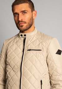 Jachetă bej matlasată cu guler în picioare, închidere cu fermoar frontal, buzunar pe piept și accente de hardware negre contrastante. Textură netedă și design ajustat.