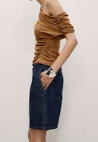 Top marrone con spalle scoperte e dettagli arricciati, abbinato a una gonna di jeans. Un bracciale a polsino d'argento indossato al polso completa l'outfit.