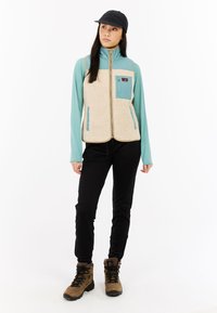Beige fleece vest met turquoise accenten, met volledige rits en twee vo pockets. Draag het over een turquoise langemouwtop en combineer het met zwarte broek en bruine laarzen.