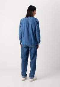Denim shirt with a back pleat, button cuffs, and a relaxed fit, paired with loose denim pants and white sneakers. All in medium blue.