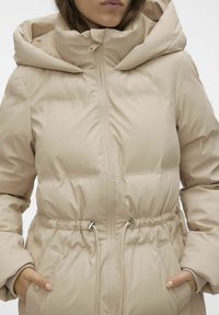 Veste beige duveteuse avec un col haut, fermeture éclair, taille élastique et poches latérales. Texture de tissu lisse et brillante.
