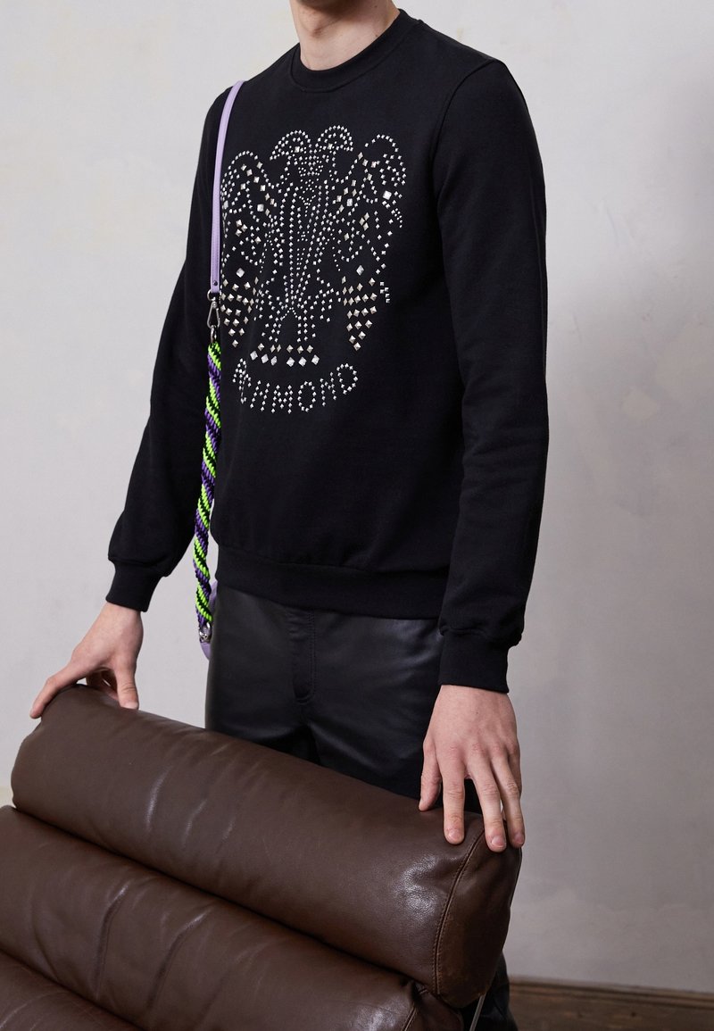 Svart sweatshirt med ett strass-eagle-mönster, bärs med läderbyxor. En flerfärgad rem korsar kroppen och vilar på en brun läderstol.