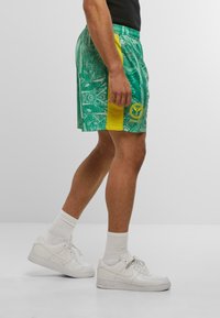 Grønne mønstrede shorts med gule sidelapper og logo, lavet af letvægtsmateriale. Sammen med hvide sneakers og ankelstrømper.