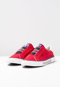 Červené plátěné slip-on boty s bílou gumovou podrážkou. Obsahují oranžové očka, vzorovaný námořnický a bílý akcentní pásek a kontrastní barevné pruhy na patě.