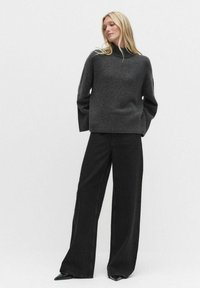 Grijze, gebreide trui met een hoge hals, gecombineerd met wijde zwarte broek. Beide kledingstukken hebben een casual pasvorm en een zachte textuur.