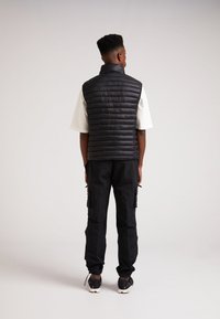 Schwarze, gepolsterte Weste mit horizontalen Nähten, hohem Kragen, getragen über einem weißen Hemd. Kombiniert mit schwarzen Cargo-Hosen und Turnschuhen.